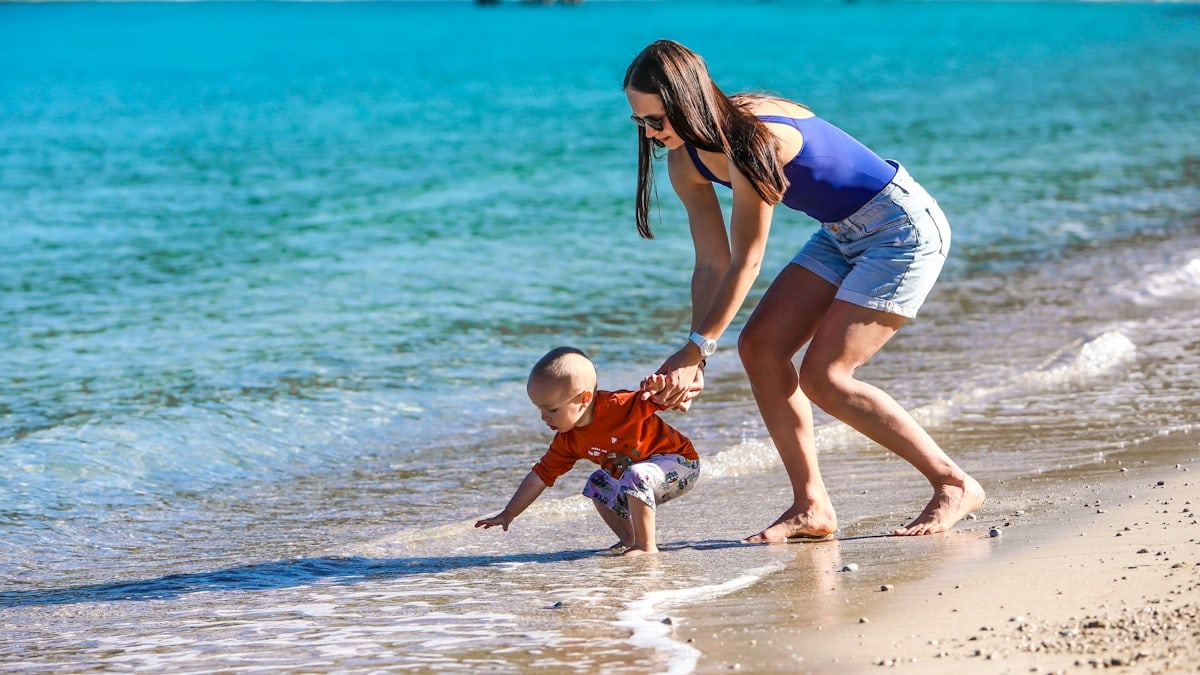woman in blue and white tank top holding baby in white onesie on beach during daytime